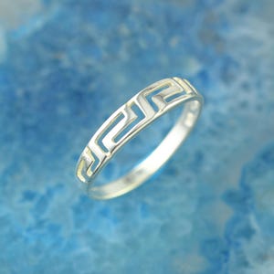 May include: A silver ring with a Greek key design. The ring features a continuous geometric pattern around the band. The ring is set against a blue background.