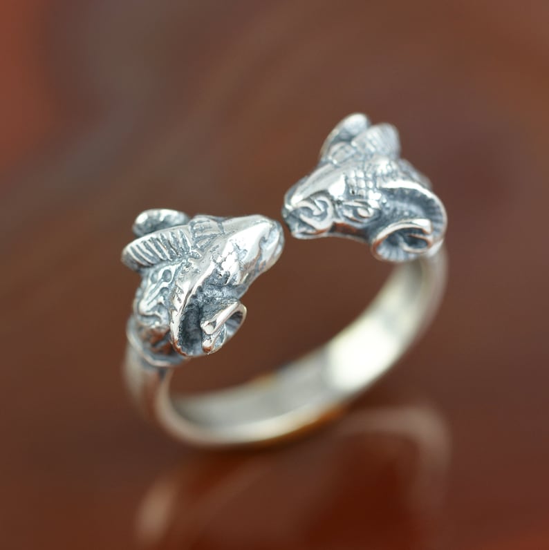 May include: A silver ring with two ram heads facing each other. The ring is open and the ram heads are detailed.