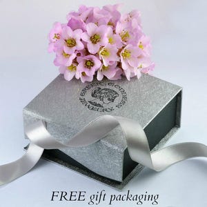 May include: A silver gift box with a black ribbon and a pink flower bouquet. The box has a logo that says "Greek Goddess Jewelry".