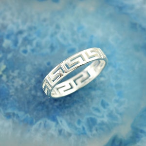 May include: A silver ring featuring a continuous cut-out Greek key pattern around the entire band. The metal has a polished, reflective finish, and the geometric design creates an openwork effect throughout the circumference of the circular jewelry piece.