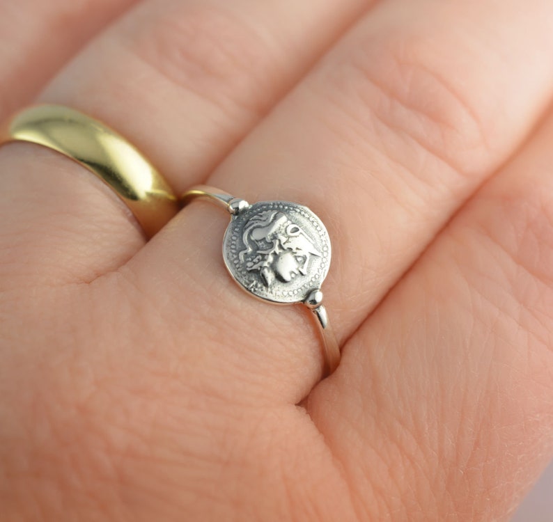 Puede incluir: Un anillo de plata con un dise&ntilde;o redondo y detallado que presenta una criatura m&iacute;tica. El anillo se lleva en un dedo.