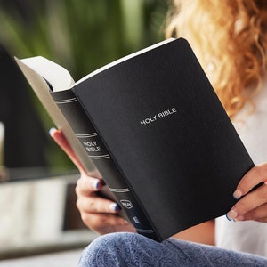 May include: A black leather-bound Holy Bible with gold lettering. The Bible is open and being read by a person.