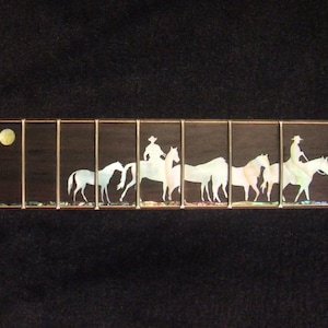 May include: A guitar neck with a desert scene inlay. The inlay features a red rock formation, cacti, a full moon, cowboys on horseback, and a dog. The fretboard is dark brown with gold frets.