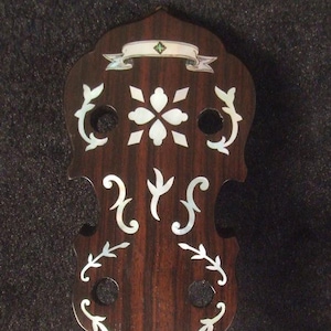 May include: Close-up of a dark wood banjo neck with a white and silver inlay design. The inlay features a banner, a flower, and scrolling vines. The text "Bruce Wei Handmade Tenor Banjo Neck BN-2015" is visible at the bottom of the image.