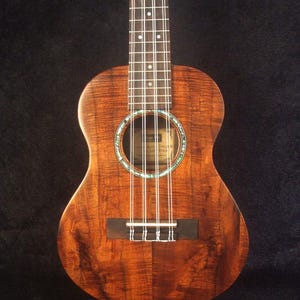May include: A tenor ukulele with eight strings, featuring a rich, brown wood grain. The sound hole is encircled with a decorative inlay. The text "Bruce Wei 8 String Tenor Ukulele UG17-3064" is visible at the top. The ukulele rests on a stand.