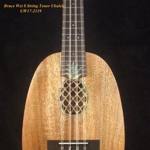 Puede incluir: Un ukelele tenor de madera marrón con ocho cuerdas y una incrustación de piña. El ukelele tiene un puente negro y una boca de sonido con un diseño de piña decorativo.