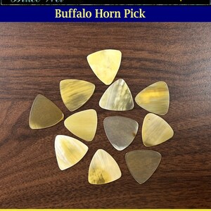 May include: A close-up of 11 buffalo horn guitar picks arranged in a circle on a brown wooden surface. The picks are all triangular in shape and have a natural, light brown color with darker brown streaks.