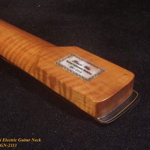 Op de afbeelding: Close-up van een houten elektrische gitaarhals. De hals heeft een lichtbruine, gevlamde esdoorn afwerking. Een rechthoekig label met tekst is ingelegd bij de kop. De basis van de hals is donkerbruin met een gouden rand.