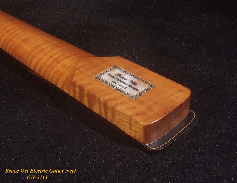 Op de afbeelding: Close-up van een houten elektrische gitaarhals. De hals heeft een lichtbruine, gevlamde esdoorn afwerking. Een rechthoekig label met tekst is ingelegd bij de kop. De basis van de hals is donkerbruin met een gouden rand.