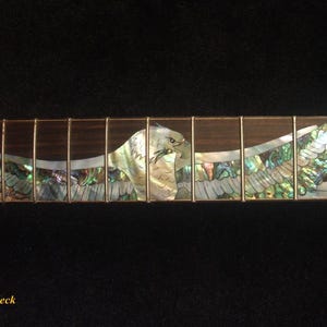 May include: Close-up of an electric guitar neck with an inlaid eagle design. The eagle is crafted from iridescent abalone shell, with a detailed depiction of the bird's head and outstretched wings. The fretboard is dark wood with metal frets.