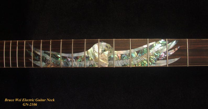 May include: Close-up of an electric guitar neck with an inlaid eagle design. The eagle is crafted from iridescent abalone shell, with a detailed depiction of the bird's head and outstretched wings. The fretboard is dark wood with metal frets.