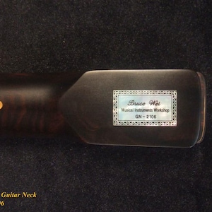 May include: Close-up of a dark brown electric guitar neck with a light-colored inlay. The headstock features a label with the text "Bruce Wei Musical Instruments Workshop GN-2106."