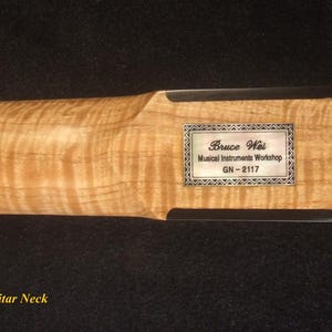 May include: Close-up of a maple electric guitar neck. The wood grain is visible. A rectangular silver label with the text "Bruce Wei Musical Instruments Workshop GN-2117" is affixed to the headstock.