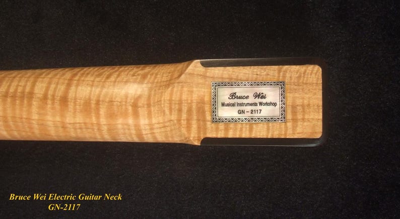 May include: Close-up of a maple electric guitar neck. The wood grain is visible. A rectangular silver label with the text "Bruce Wei Musical Instruments Workshop GN-2117" is affixed to the headstock.