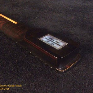 May include: Close-up of a dark brown electric guitar neck with a light-colored stripe down the center. A rectangular inlay with text is visible on the headstock. The neck is made of wood and has a glossy finish.