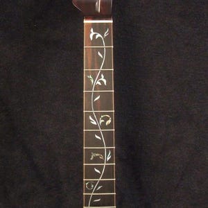 May include: A dark brown electric guitar neck featuring a headstock with six tuning peg holes and a fretboard inlaid with a continuous mother-of-pearl vine pattern. The text "Bruce Wei Electric Guitar Neck GN-2127" is printed on the left side.