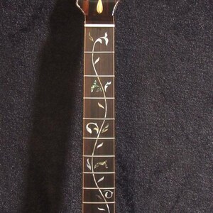 May include: A dark brown electric guitar neck with a decorative floral inlay. The headstock has tuning peg holes and a mother-of-pearl inlay. The text "Bruce Wei Electric Guitar Neck GN-2113" is visible.