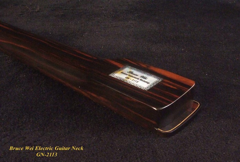 May include: A close-up of a Bruce Wei electric guitar neck, model GN-2113. The neck is made of dark wood with reddish-brown and black grain patterns. A rectangular label with text is visible.
