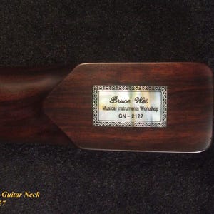 May include: A dark brown wooden electric guitar neck featuring a rectangular mother-of-pearl inlay on the heel. The inlay is engraved with the text Bruce Wei, Musical Instruments Workshop, and the model number GN-2127 in a decorative border.