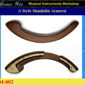 May include: A wooden mandolin armrest with a curved shape and a brown finish. The armrest is designed to be attached to a mandolin, providing support for the player's arm. The armrest is shown in two pieces, with adhesive tape on the back.