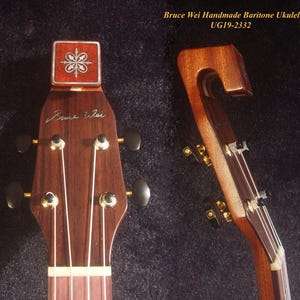May include: Close-up of a handmade baritone ukulele. The wooden instrument features a decorative headstock with a floral design and the inscription "Bruce Wei." The strings and tuning pegs are visible.