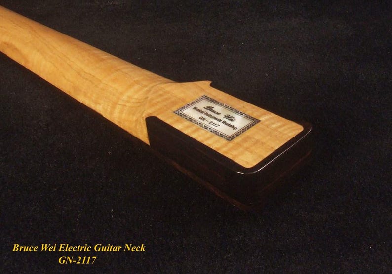 May include: Close-up of a maple electric guitar neck with a dark brown heel. The neck has a light-colored, patterned wood grain. A rectangular label with text is affixed to the heel. The text at the bottom reads "Bruce Wei Electric Guitar Neck GN-2117".
