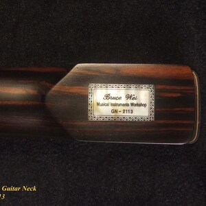 May include: A close-up of a dark brown electric guitar neck with a rectangular inlay. The inlay reads "Bruce Wei Musical Instruments Workshop GN-2113" within a decorative frame. The wood grain is visible.