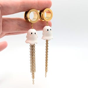 Könnte beinhalten: Ein Paar goldfarbene Ohr-Dehnstäbe mit weißen Geisteranhängern und baumelnden Goldketten. Die Geisteranhänger haben schwarze Augen und einen lächelnden Ausdruck. Der Hintergrund ist weiß.