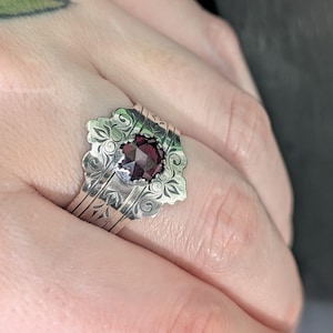 May include: A silver ring featuring a floral design and a faceted, deep red gemstone. The ring has an ornate, scalloped setting and engraved details on the band. The gemstone is the focal point, set in a detailed, decorative frame.