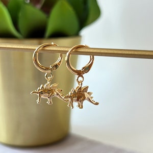 Op de afbeelding: Gouden creolen met een hangende dinosaurusbedel. De dinosaurus is goudkleurig en heeft een getextureerd oppervlak.