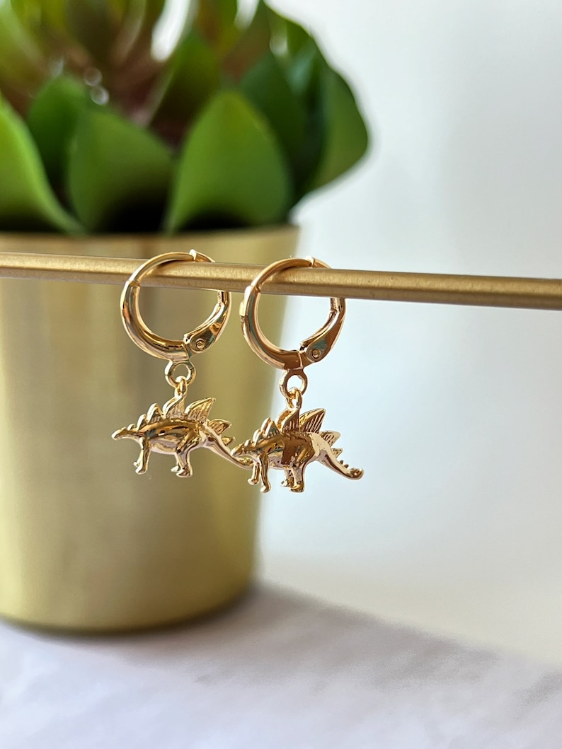 Op de afbeelding: Gouden creolen met een hangende dinosaurusbedel. De dinosaurus is goudkleurig en heeft een getextureerd oppervlak.
