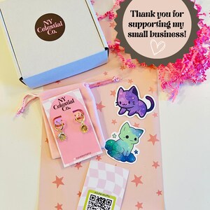 Op de afbeelding: Een roze en witte cadeauzak met een witte doos, een klein zakje met gouden oorbellen en twee stickers met cartoonkatten. De tas heeft een roze en wit sterrenpatroon en de tekst "Bedankt voor het steunen van mijn kleine bedrijf!" op een bruine stervormige sticker. De tas heeft ook een QR-code sticker met de tekst "cute stuffa".