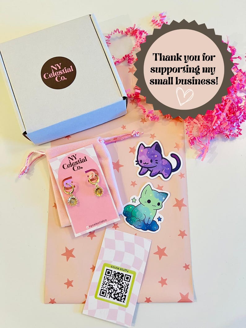 Op de afbeelding: Een roze en witte cadeauzak met een witte doos, een klein zakje met gouden oorbellen en twee stickers met cartoonkatten. De tas heeft een roze en wit sterrenpatroon en de tekst "Bedankt voor het steunen van mijn kleine bedrijf!" op een bruine stervormige sticker. De tas heeft ook een QR-code sticker met de tekst "cute stuffa".