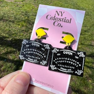 May include: A pair of black and white Ouija board earrings with a yellow lemon charm. The earrings are on a pink card with the text "NY Celestial Co."