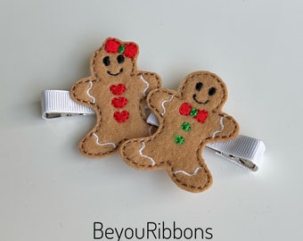 May include: Two felt gingerbread people hair clips. The gingerbread person on the left has a red bow and three red hearts on the front. The gingerbread person on the right has a red bow tie and two green dots on the front. The text "BeyouRibbons" is printed below the gingerbread people.