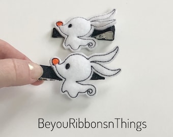 May include: Two white felt hair clips with black stitching and orange noses. The clips are shaped like a cartoon ghost with a single eye and a small smile. The clips are attached to black alligator clips.