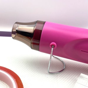 May include: A pink heat gun with a brown nozzle and a metal stand. The heat gun is on a white surface.