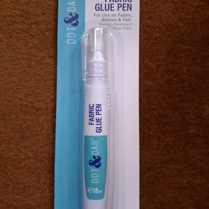 May include: A Fabric Glue Pen by Dot & Dab, in a clear plastic blister pack. The pen is white and blue, and is labelled "Fabric Glue Pen". It is designed for fabric, ribbon, and felt. The packaging states it is strong, permanent, and dries clear. Contains 18ml.