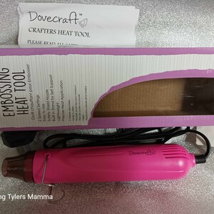May include: Pink Dovecraft embossing heat tool with a black cord and a clear nozzle. The tool has two speed settings and a safety shield nozzle. The packaging says "Embossing Heat Tool" and "Outil chauffant pour Embosser".