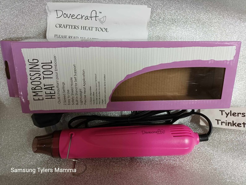 May include: Pink Dovecraft embossing heat tool with a black cord and a clear nozzle. The tool has two speed settings and a safety shield nozzle. The packaging says "Embossing Heat Tool" and "Outil chauffant pour Embosser".