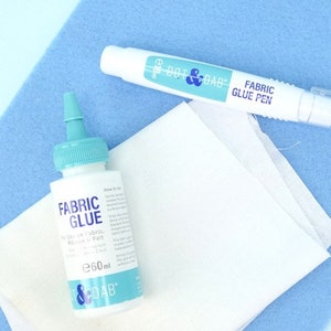 May include: A white bottle of fabric glue with a blue cap and a white fabric glue pen with a blue cap. Both products are labeled "Dot & Dab" and are on a blue surface.