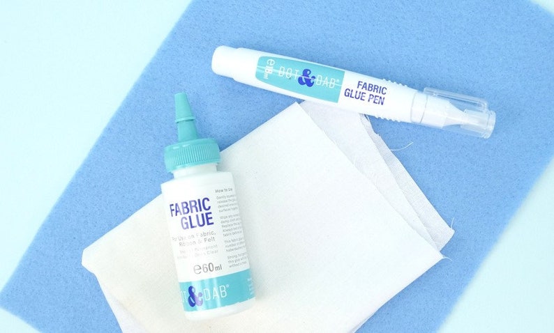 May include: A white bottle of fabric glue with a blue cap and a white fabric glue pen with a blue cap. Both products are labeled "Dot & Dab" and are on a blue surface.