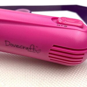 May include: A pink Dovecraft Essentials heat gun with a black on/off switch labeled "0/1/2". The heat gun has a vent with five slots and a small bird logo.
