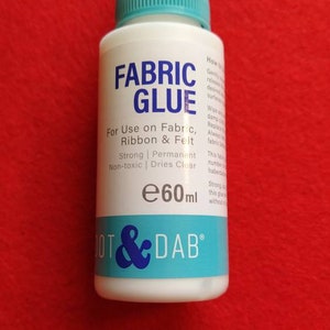 May include: A white bottle of Dot & Dab Fabric Glue with a blue cap and label. The label says "Fabric Glue" and "For Use on Fabric, Ribbon & Felt". It also says "Strong | Permanent | Non-toxic | Dries Clear" and "e60ml".