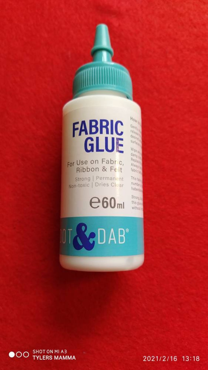 May include: A white bottle of Dot & Dab Fabric Glue with a blue cap and label. The label says "Fabric Glue" and "For Use on Fabric, Ribbon & Felt". It also says "Strong | Permanent | Non-toxic | Dries Clear" and "e60ml".