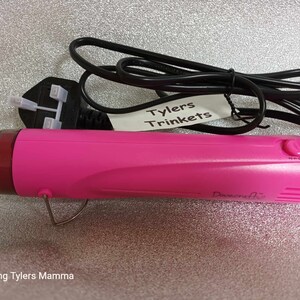 May include: A pink heat gun with a black power cord and a black plug. The heat gun has a black switch with the letters "NPO" on it. The heat gun is labeled "Dovecrafts". A white label with the text "Tylers Trinkets" is attached to the cord.