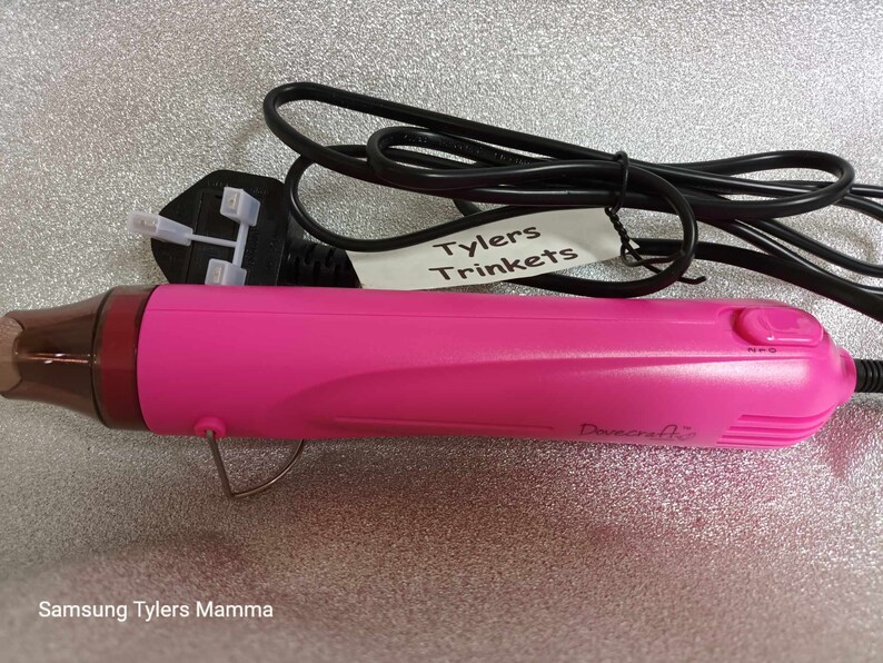 May include: A pink heat gun with a black power cord and a black plug. The heat gun has a black switch with the letters "NPO" on it. The heat gun is labeled "Dovecrafts". A white label with the text "Tylers Trinkets" is attached to the cord.