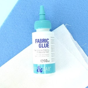 May include: A clear plastic bottle of fabric glue with a blue cap and label. The label reads "FABRIC GLUE" and "For Use on Fabric, Ribbon & Felt". The bottle is 60 ml and is labeled "DOT & DAB".