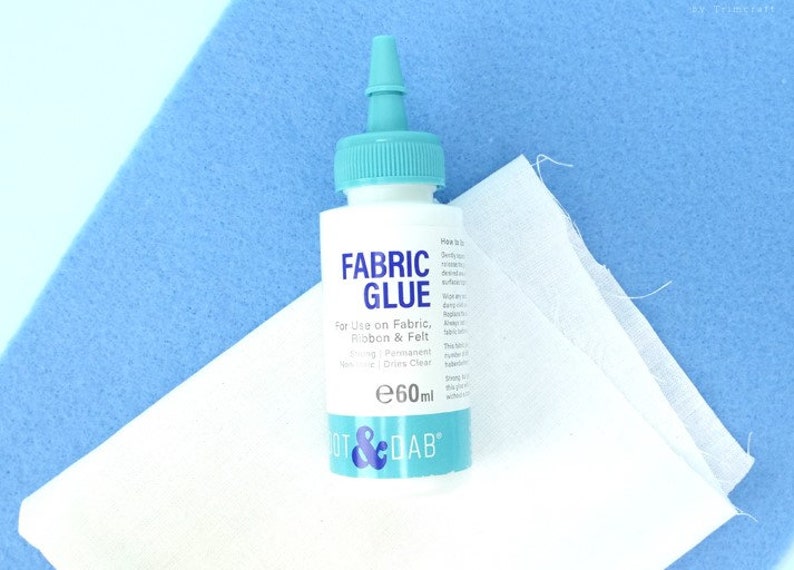 May include: A clear plastic bottle of fabric glue with a blue cap and label. The label reads "FABRIC GLUE" and "For Use on Fabric, Ribbon & Felt". The bottle is 60 ml and is labeled "DOT & DAB".