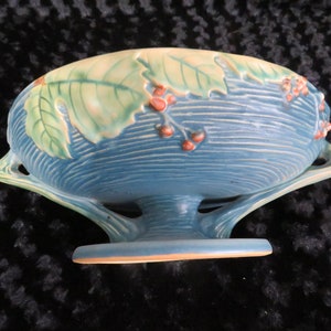 May include: A blue and green ceramic bowl with a leaf and berry design. The bowl has two handles and a pedestal base.